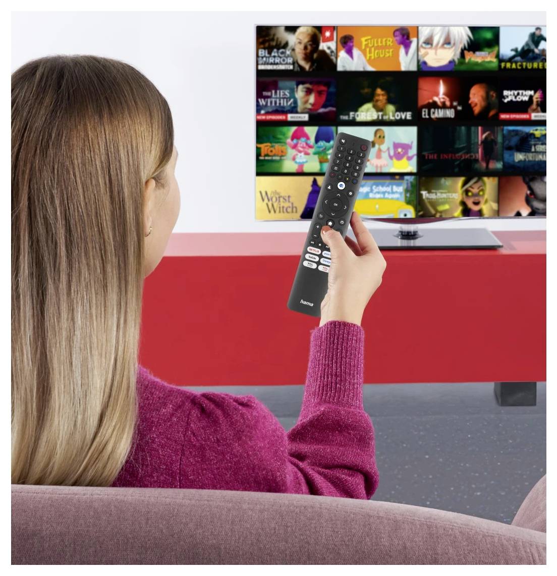 Frau sitzt auf einem Sofa und zeigt mit einer Fernbedienung auf einen Fernseher, der ein Streaming-App-Menü mit Film- und Serien-Miniaturansichten anzeigt.