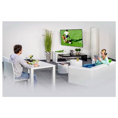 Zwei Menschen entspannen sich in einem Wohnzimmer und schauen sich ein Fußballspiel auf einem an der Wand montierten Fernseher an, wobei einer an einem Tisch sitzt und der andere auf einem Sofa liegt und ein Tablet nutzt.