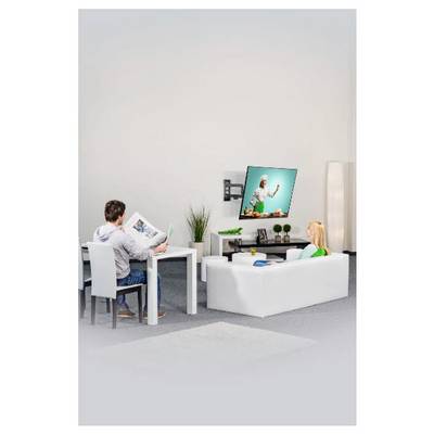 Ein Mann liest eine Zeitschrift an einem Tisch, während ein Kind auf einem Sofa sitzt und im hellen Wohnzimmer Fernsehen schaut.