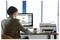 Eine Person, die an einem Desktop-Computer neben einem Drucker in einem hellen Büro arbeitet, von hinten betrachtet.