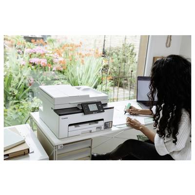 Frau arbeitet an einem Schreibtisch neben einem Canon-Drucker und schreibt auf Papier in einem Heimbüro mit einem Laptop und Blick auf den Garten.