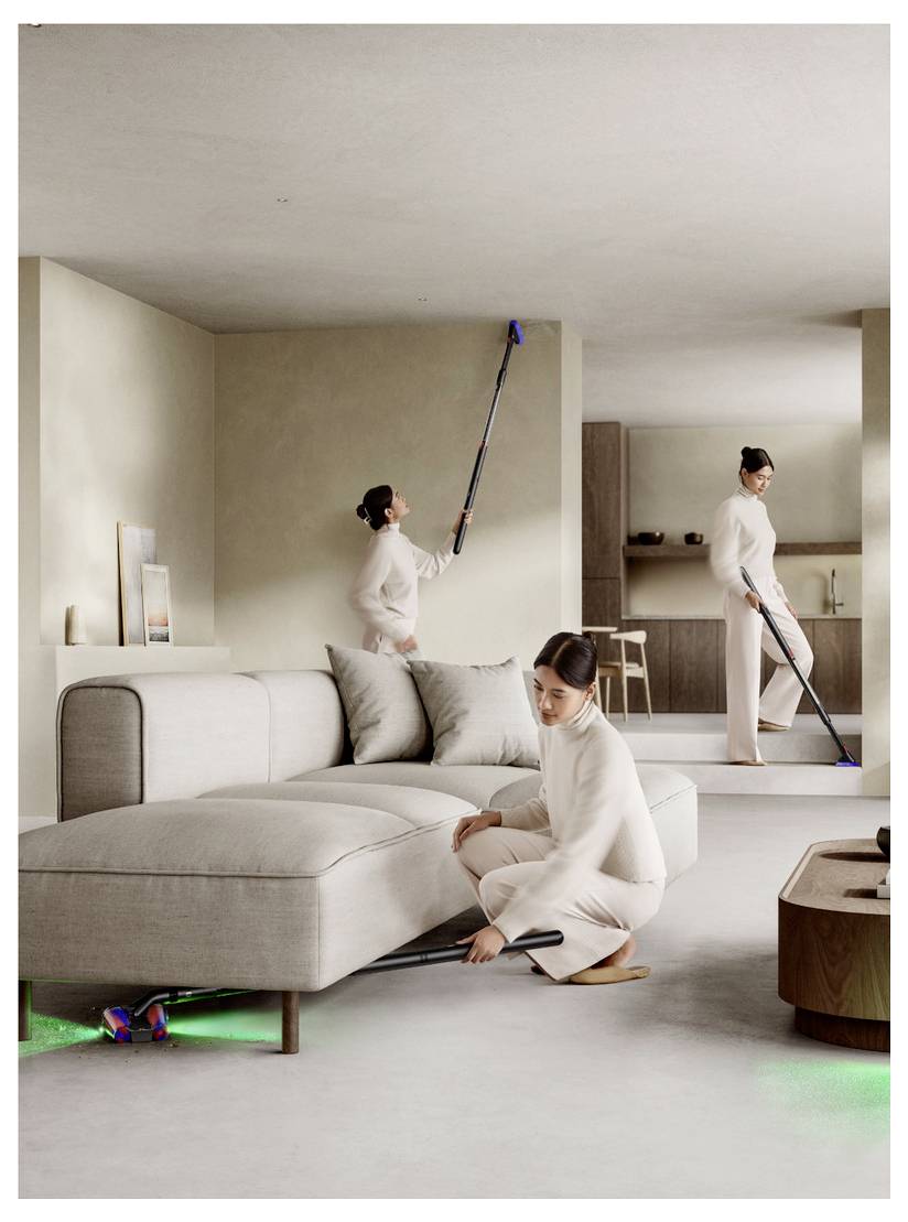 Drei Frauen in Weiß reinigen ein modernes beiges Wohnzimmer, wobei sie Wischmöpel, einen Staubwedel und einen Staubsauger unter dem Sofa und entlang der Wand verwenden.