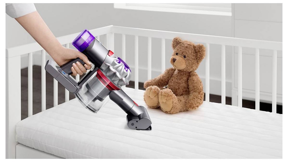 Eine Hand benutzt einen kabellosen Stabstaubsauger, um eine weiße Krippe Matratze neben einem Teddybär zu reinigen, was auf Nursery-Reinigung hindeutet.