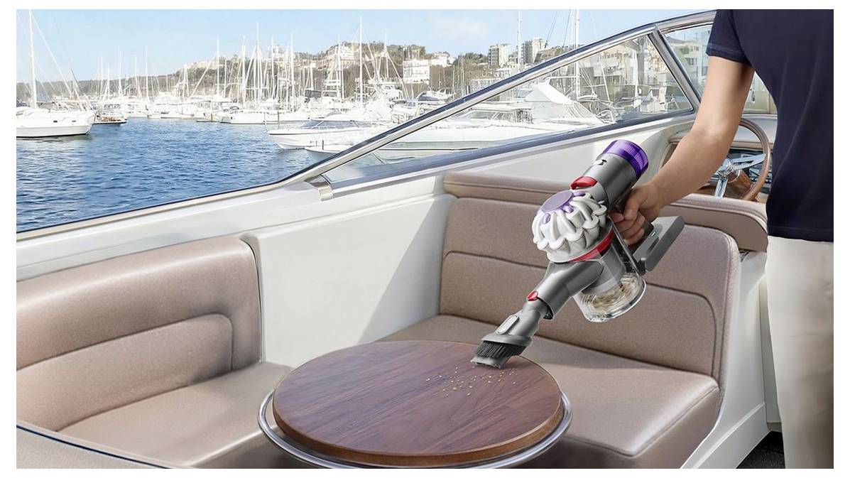Person benutzt einen kabellosen Staubsauger, um Krümel vom Tisch in der Kabine eines Bootes zu reinigen, wobei ein Jachthafen und Segelboote draußen sichtbar sind.
