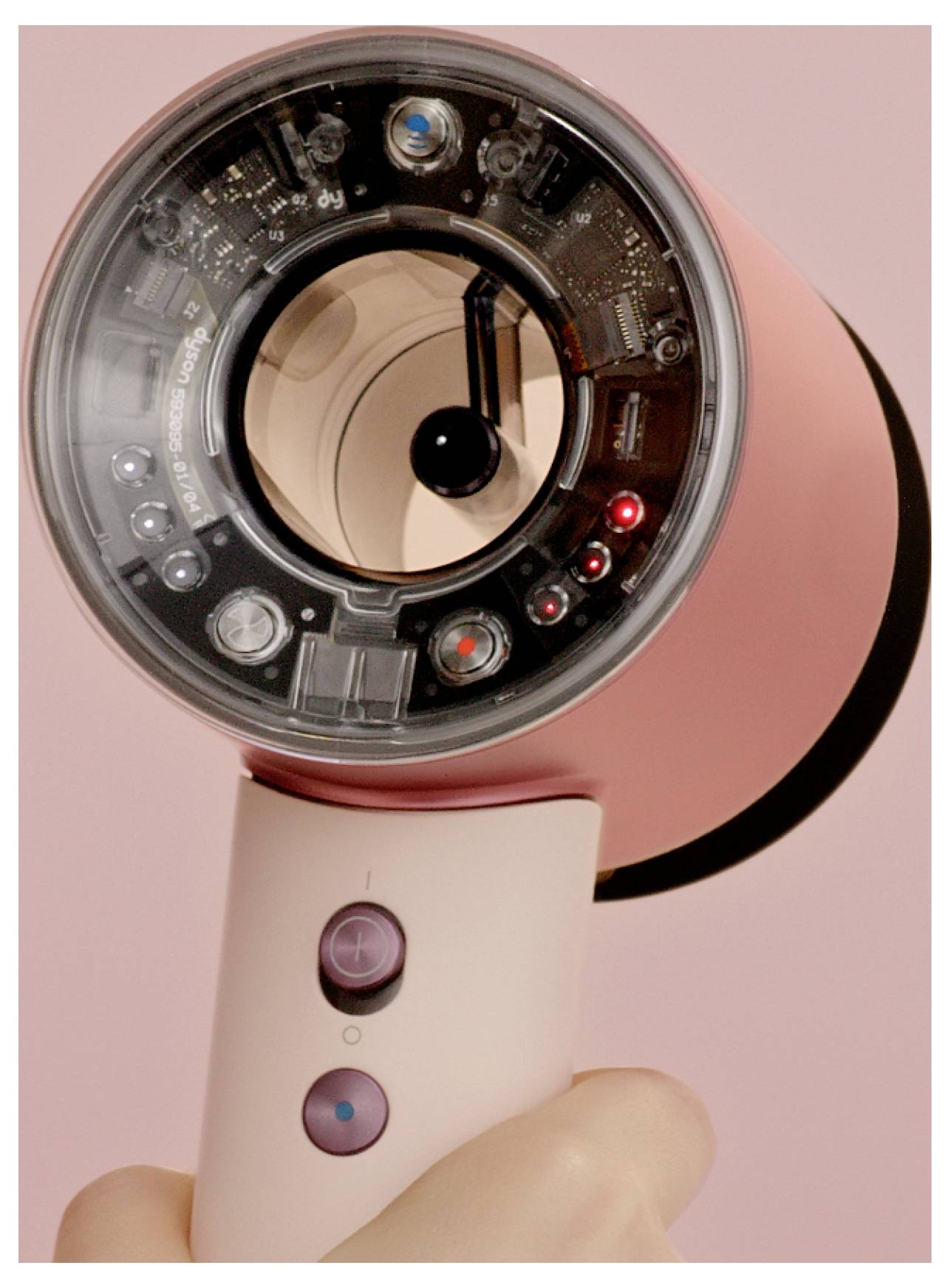 Nahaufnahme eines rosa tragbaren elektronischen Geräts mit kreisförmiger transparenter Linse/Display und leuchtenden Anzeigelichtern, das eine Hand gegen einen rosa Hintergrund hält.
