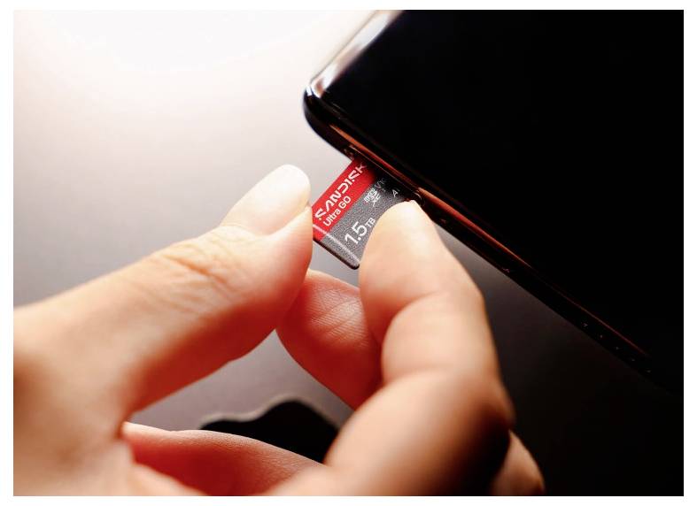 Hände führen eine mit „SanDisk" gekennzeichnete microSD-Karte in den Speicherkartensteckplatz eines Smartphones ein.