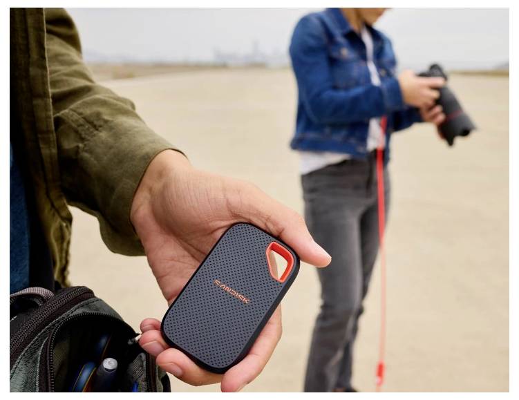Nahaufnahme einer Hand, die ein kleines tragbares schwarzes Gerät mit orangefarbenen Akzenten hält, während eine Person im Hintergrund eine Kamera am Strand hält.