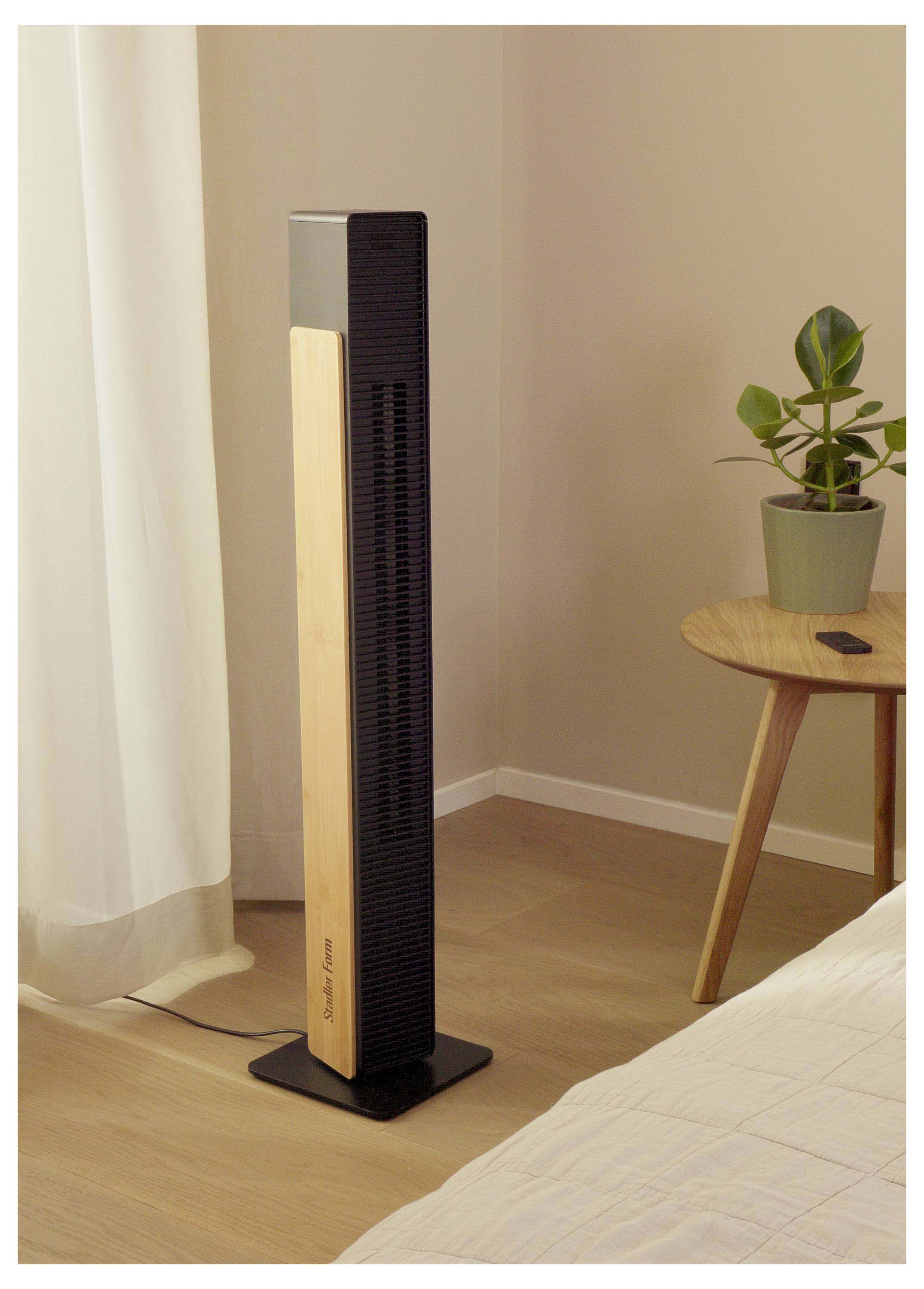 Ein hoher schwarz-weiß Turmventilator steht auf dem Boden in einem Schlafzimmer, neben einem kleinen Tisch mit einer Topfpflanze und in der Nähe eines Bettes und eines Vorhangs.