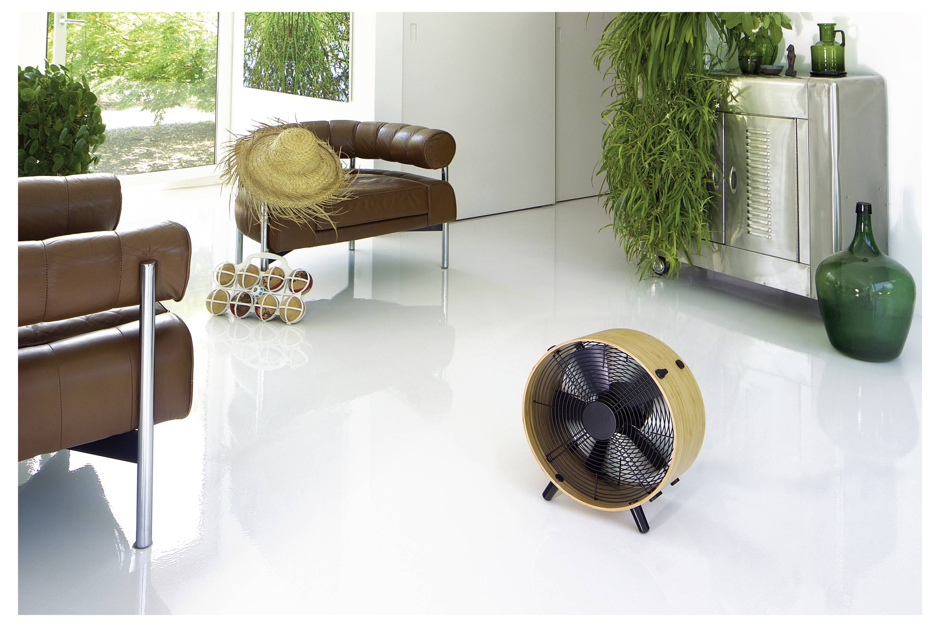 Standventilator in einem hellen, modernen Wohnzimmer mit Lederstühlen, einem Strohhut und einer grünen Vase neben einem wandmontierten Heizgerät und Pflanzen.