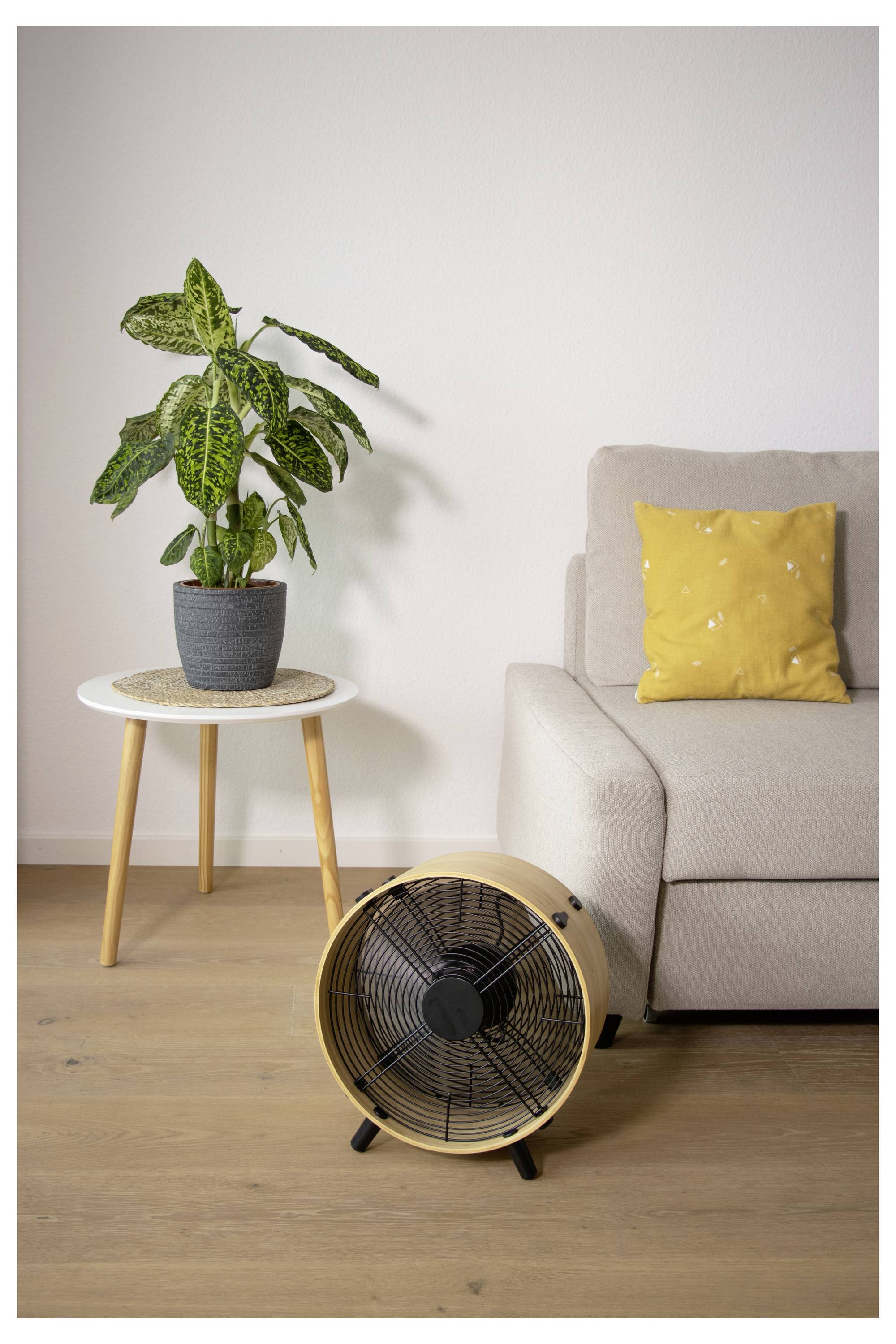 Standventilator vor einem beigefarbenen Sofa und gelbem Kissen, neben einer Topfpflanze auf einem kleinen Tisch in einem minimalistischen Wohnzimmer.