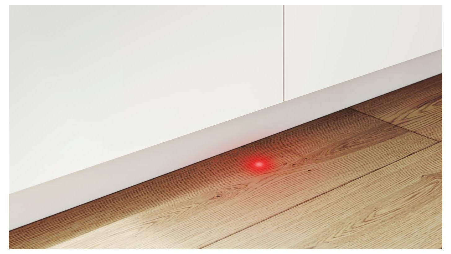 Roter Laserpunkt auf einem Holzboden neben einem weißen Schrank, der einen Sensor oder Ausrichtungspunkt andeutet.