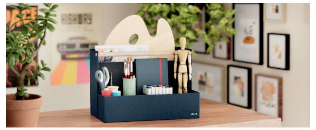 Schreibtischorganizer-Box mit Kunstmaterialien, Scheren, Markern und einer hölzernen Gliederpuppe auf einem Holztisch in einem Studio-Arbeitsbereich.