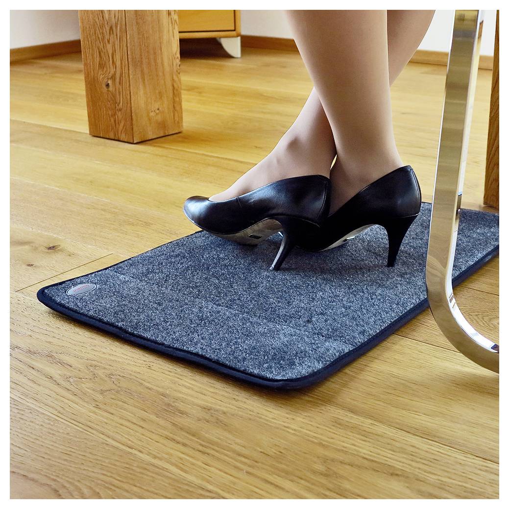 Gekreuzte Beine einer Frau in schwarzen hochhackigen Schuhen, die auf einer blauen Antiermüdungsmatte auf einem Holzboden neben einem Tisch stehen.