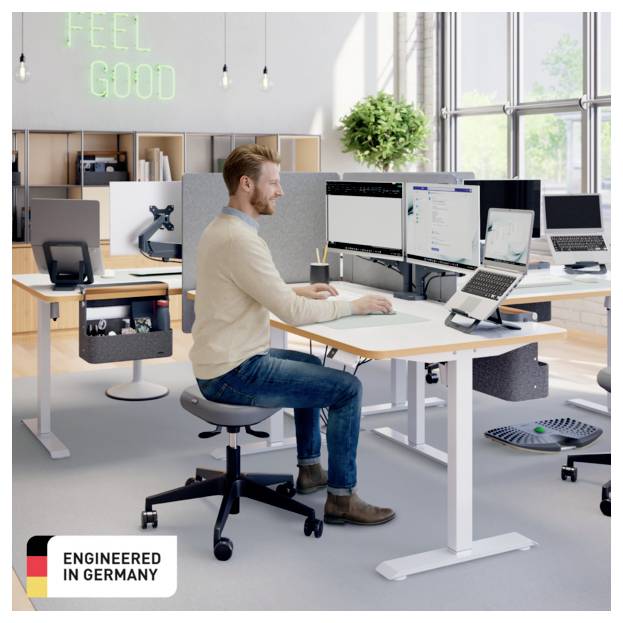 Ein Mann arbeitet an einem Stehpult in einem modernen Büro mit mehreren Monitoren; sein Ausweis zeigt 'ENGINEERED IN GERMANY' und ein Neonschild sagt 'FEEL GOOD'.