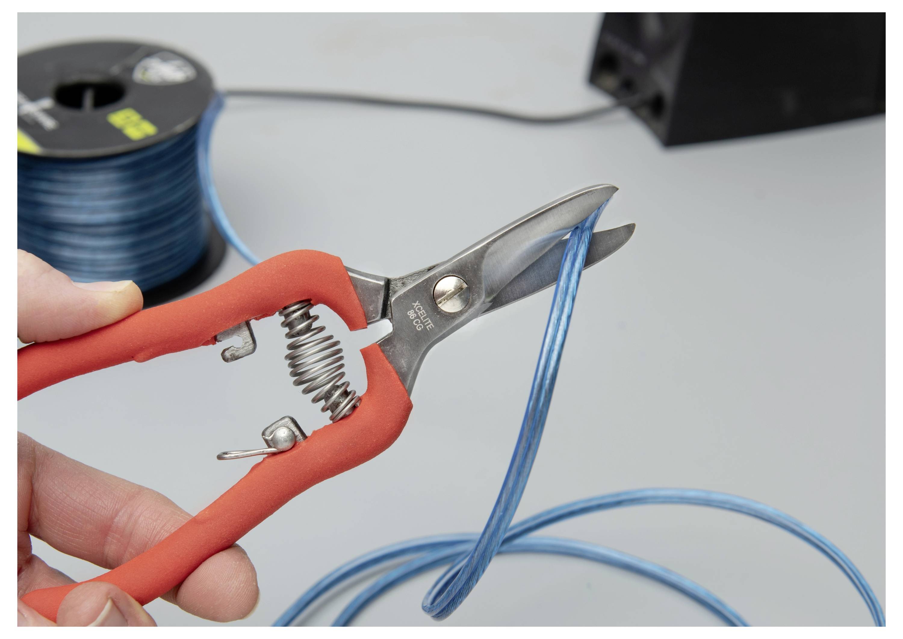 Hand, die rote Drahtschneider benutzt, um ein blaues Kabel auf einer Werkbank zu schneiden, mit einer Drahtspule im Hintergrund.