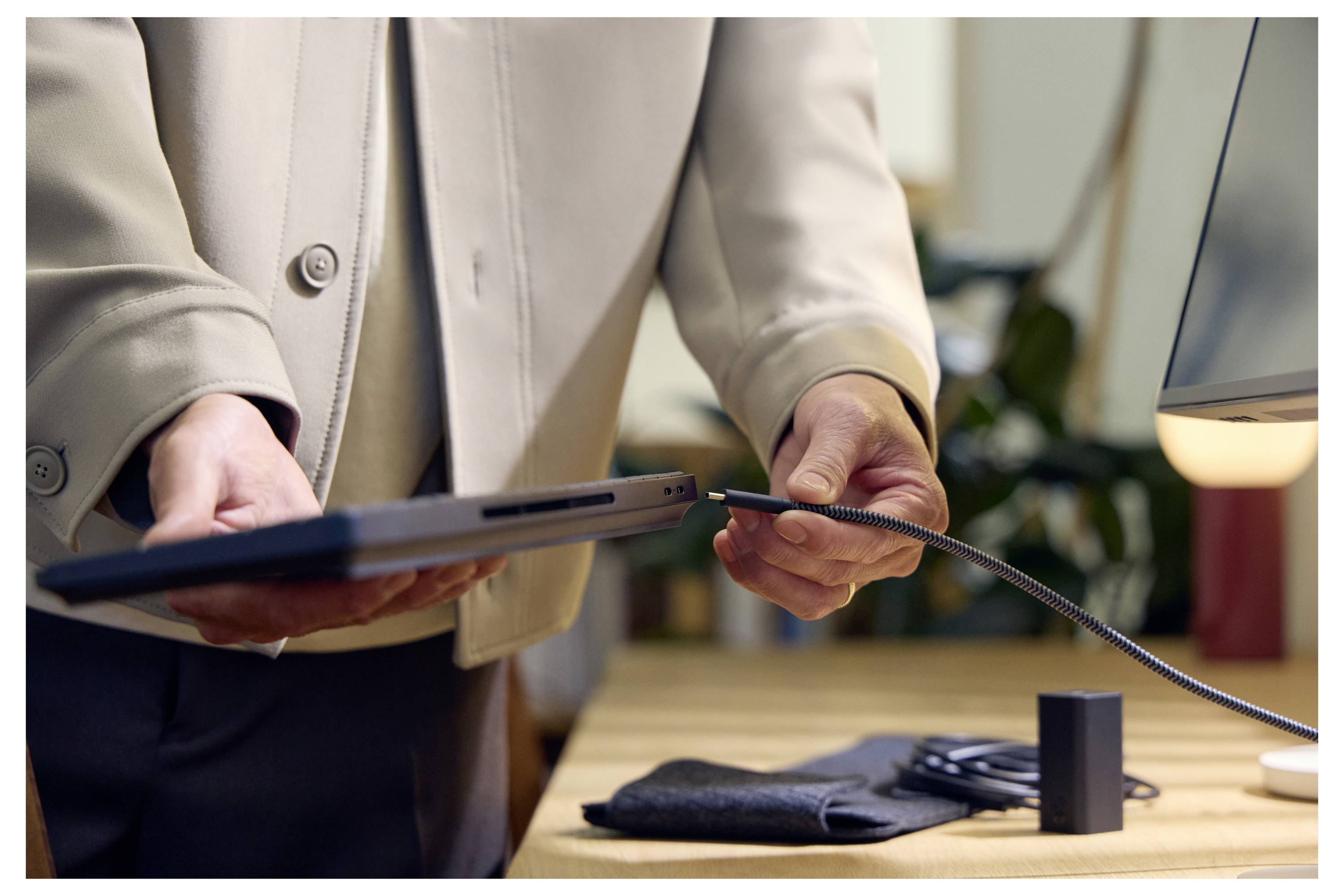 Eine Person verbindet ein Kabel mit einem Laptop an einem Schreibtisch, neben einer Lampe und kleinen Bürogegenständen.