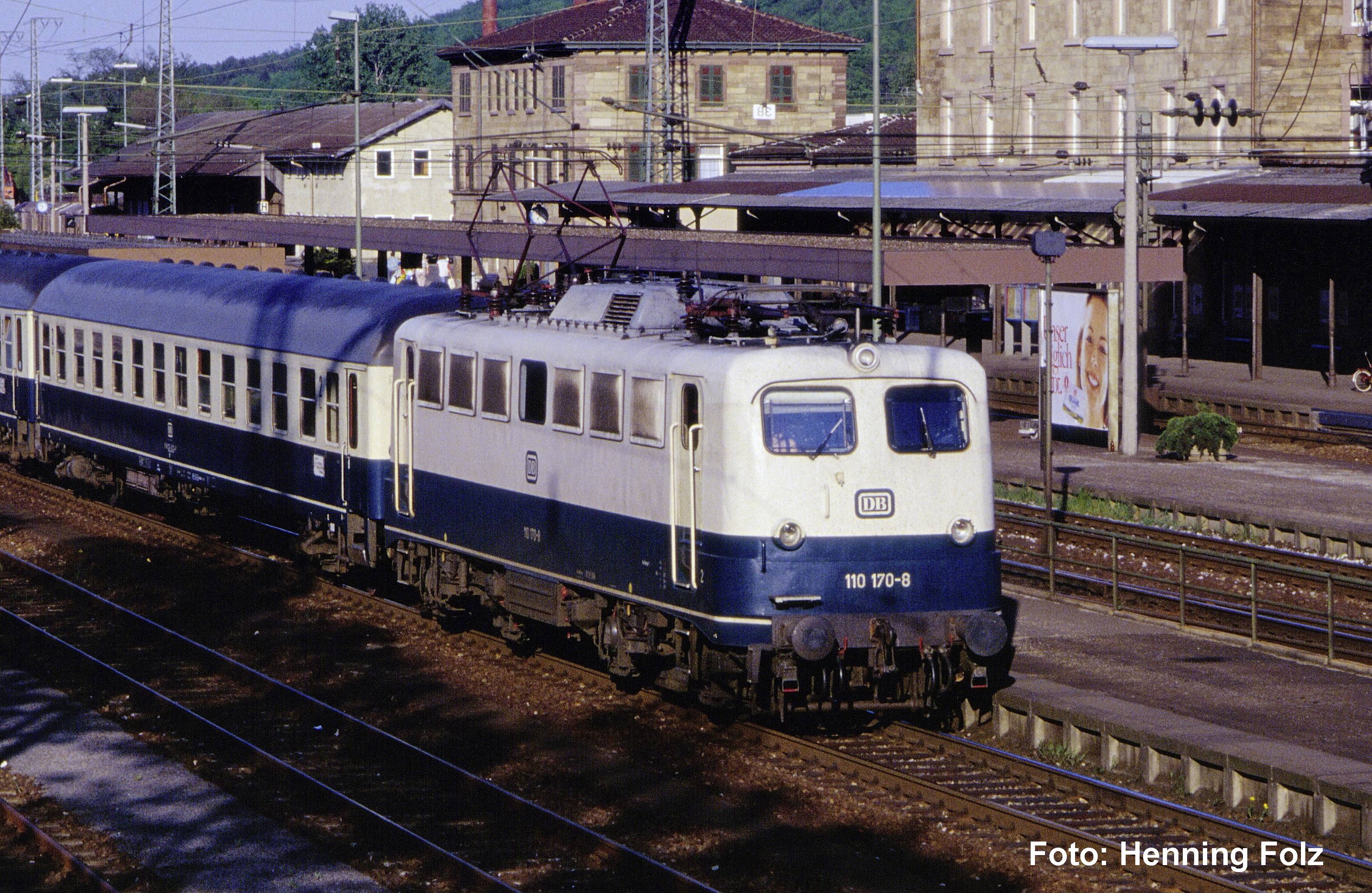 Blau-weißer elektrischer Personenzug an einem Bahnsteig, mit Fahrleitungen und Bahnhofsgebäuden im Hintergrund.