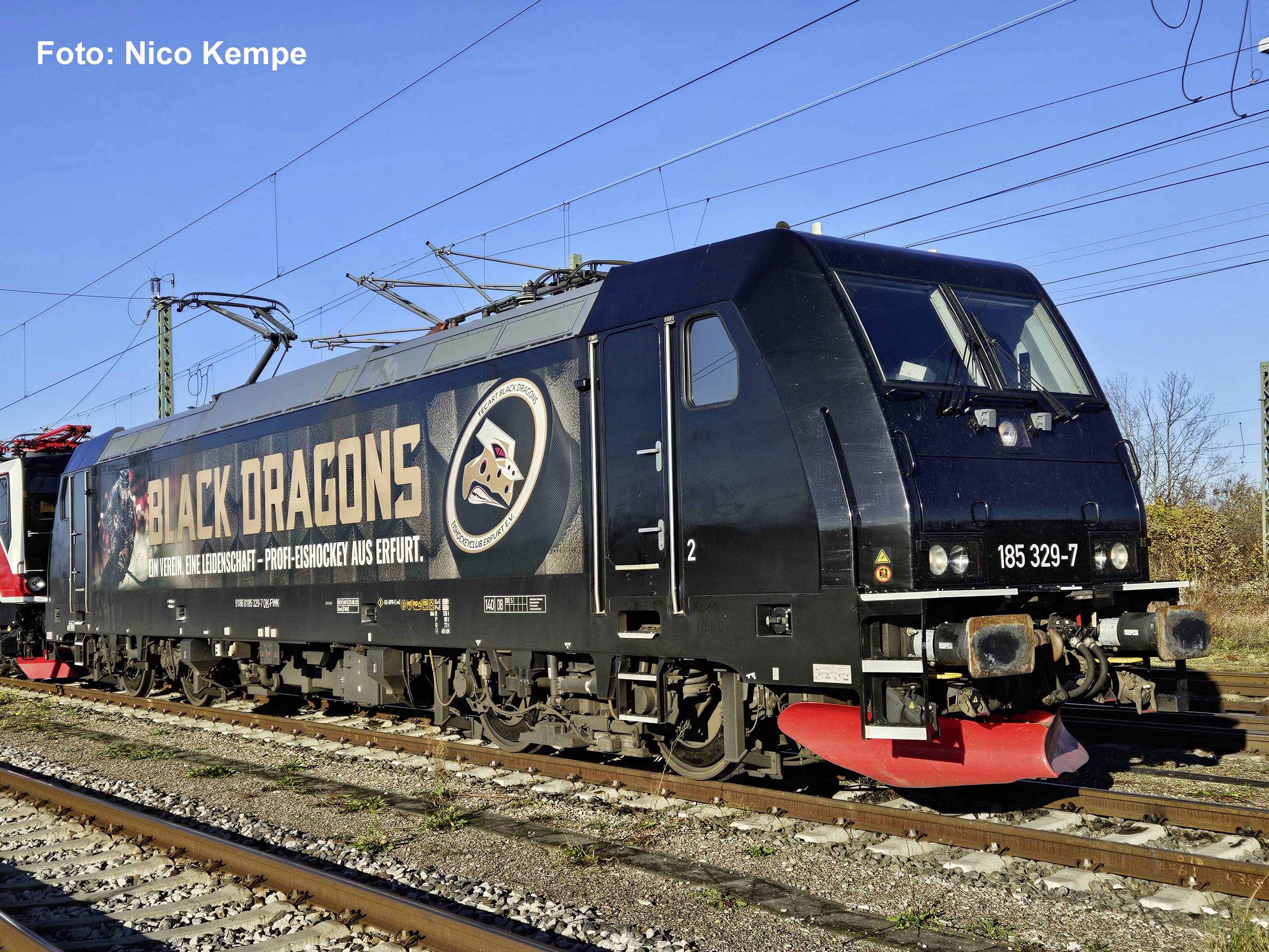 Schwarze elektrische Lokomotive mit der Aufschrift „BLACK DRAGONS" auf Eisenbahnschienen unter Oberleitung, mit einer roten Schneepflug und großem Logo an der Seite.