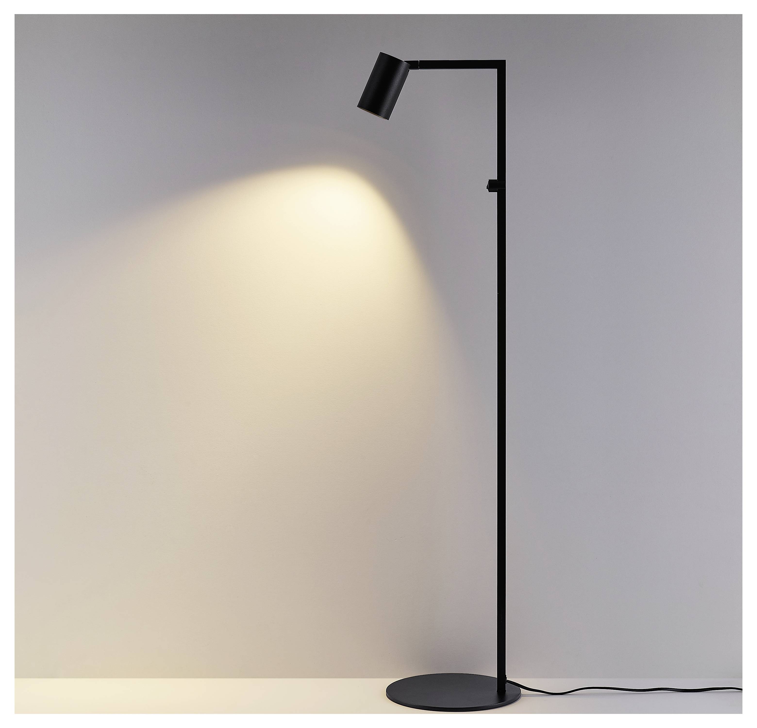 Schwarze Stehleuchte mit fokussiertem Scheinwerfer, der einen warmen Lichtkreis auf eine schlichte Wand wirft, Kabel an der Basis sichtbar.