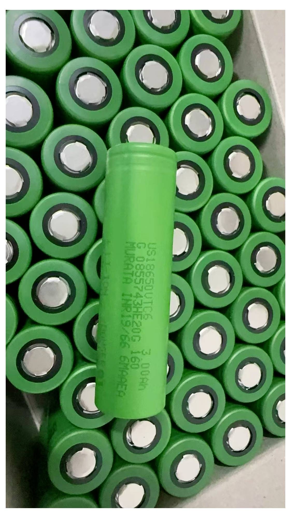 Nahaufnahme von grünen zylindrischen Batterien, die in einem Tablett verpackt sind, mit einer Batterie in der Mitte, die aufrecht steht und gedruckte Spezifikationen zeigt.