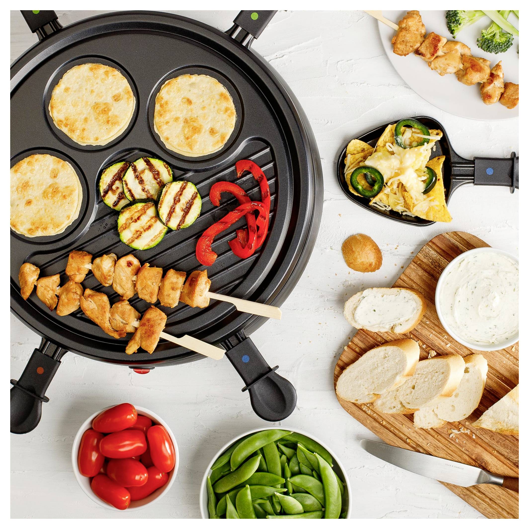 Draufsicht auf ein Tisch-Grillbrett mit Hühncherspießen, Fladen­broten, gegrilltem Gemüse, Tomaten, Zuckerschoten, Brot und Dip­sauce.