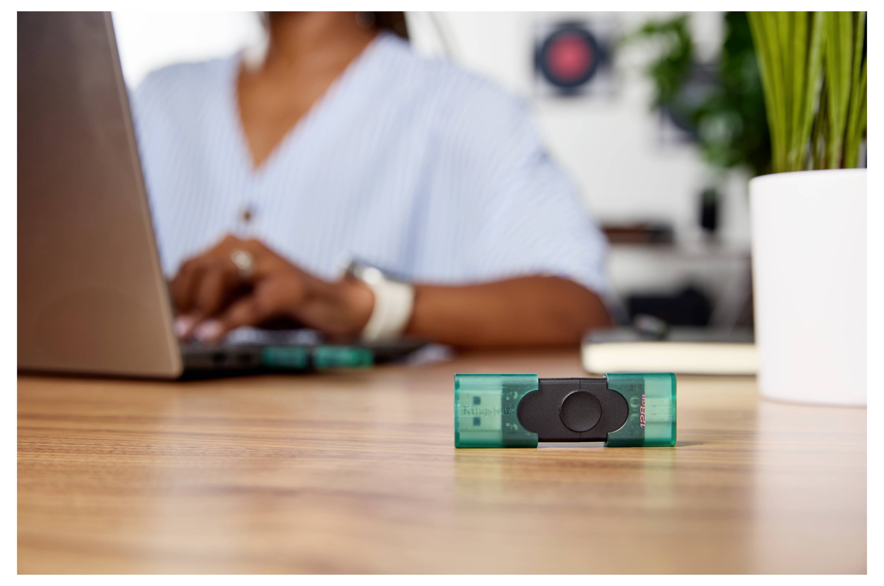Nahaufnahme eines USB-Sticks auf einem Schreibtisch, mit einer unscharfen Person, die im Hintergrund auf einem Laptop tippt.