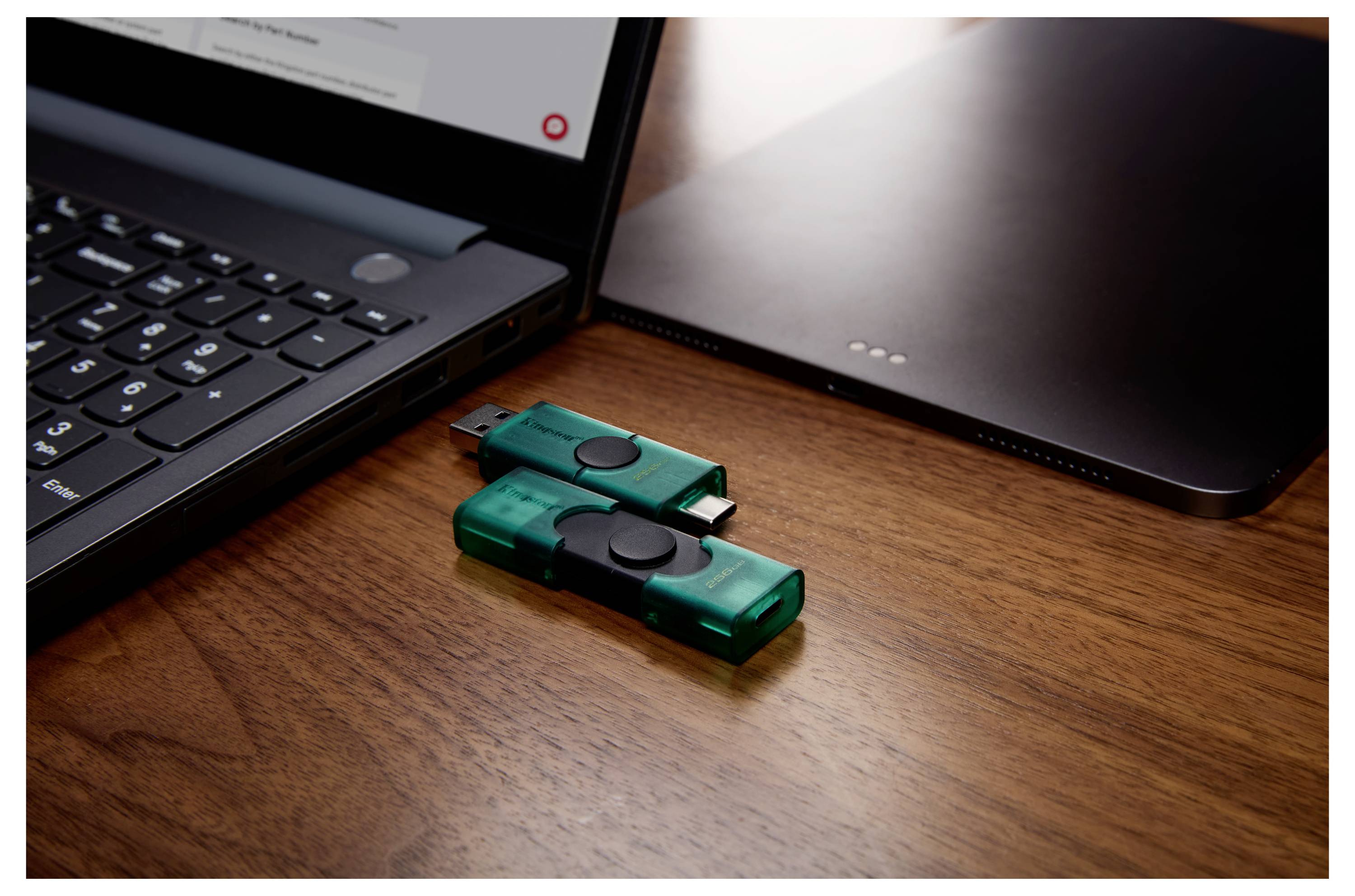 Zwei grüne USB-Speichersticks auf einem Holzschreibtisch neben einem Laptop und einem geschlossenen Gerät deuten auf Dateispeicherung oder -übertragung hin.