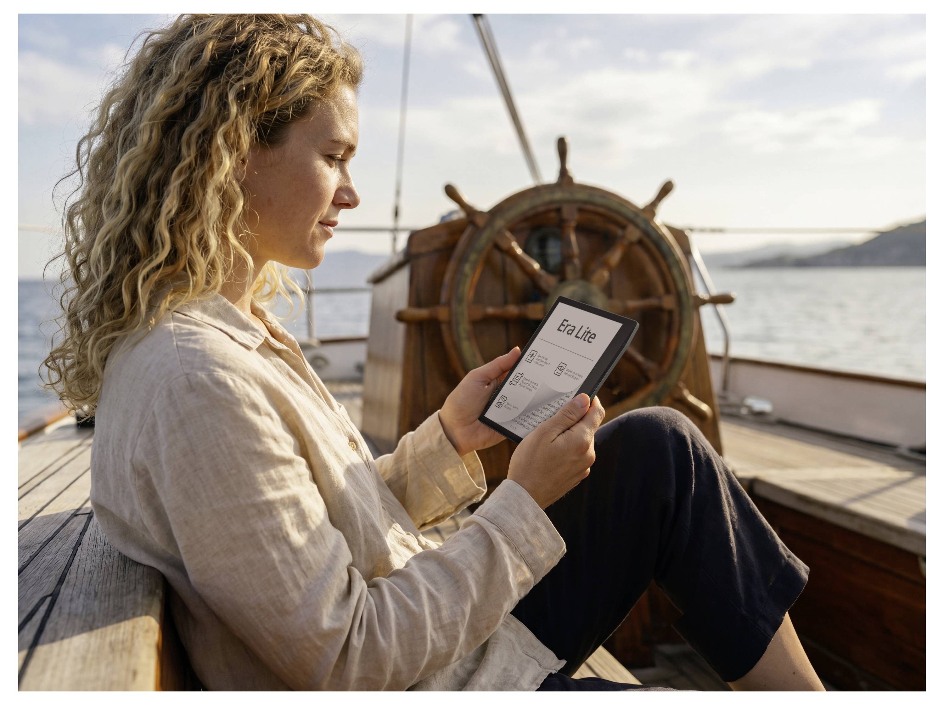 Frau sitzt auf dem Deck eines Segelboots und liest auf einem E-Reader, mit dem Steuerrad und ruhigem Wasser im Hintergrund.