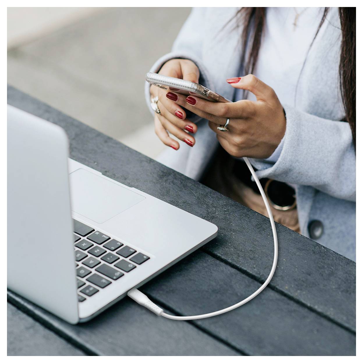 Eine Person, die ein Smartphone verwendet, das durch ein Kabel mit einem Laptop auf einem Tisch verbunden ist, was auf Datenübertragung oder Laden hindeutet.
