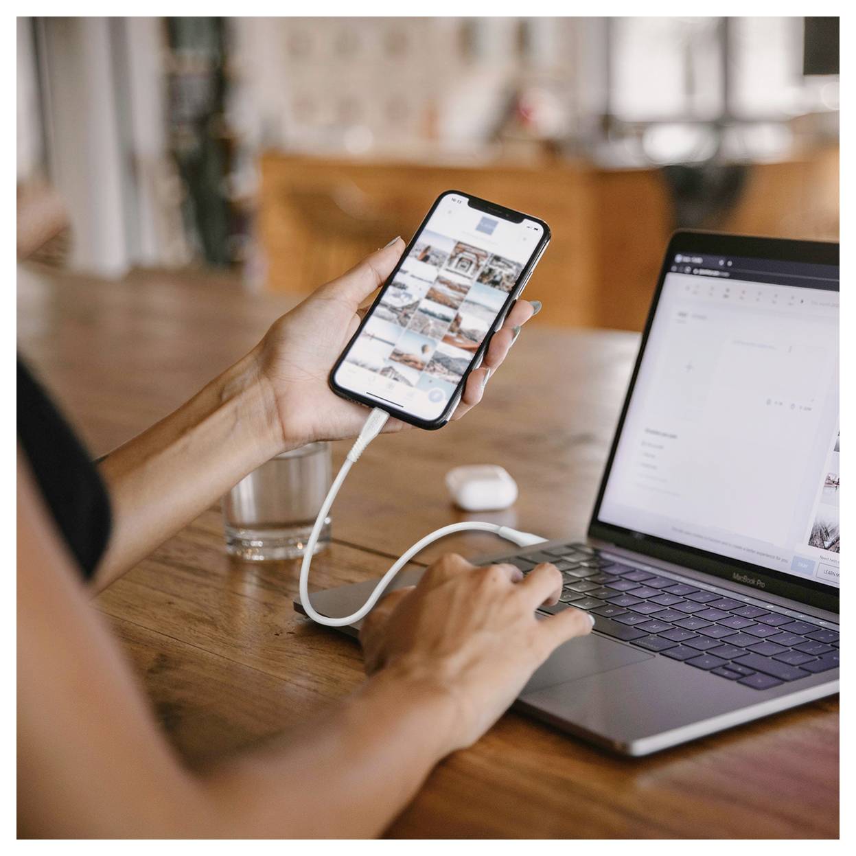 Eine Person nutzt ein Smartphone, das über ein Kabel mit einem Laptop verbunden ist, während sie an einem Holztisch arbeitet. Das Telefon zeigt eine Fotogalerie und der Laptop zeigt einen Dokumenteneditor.