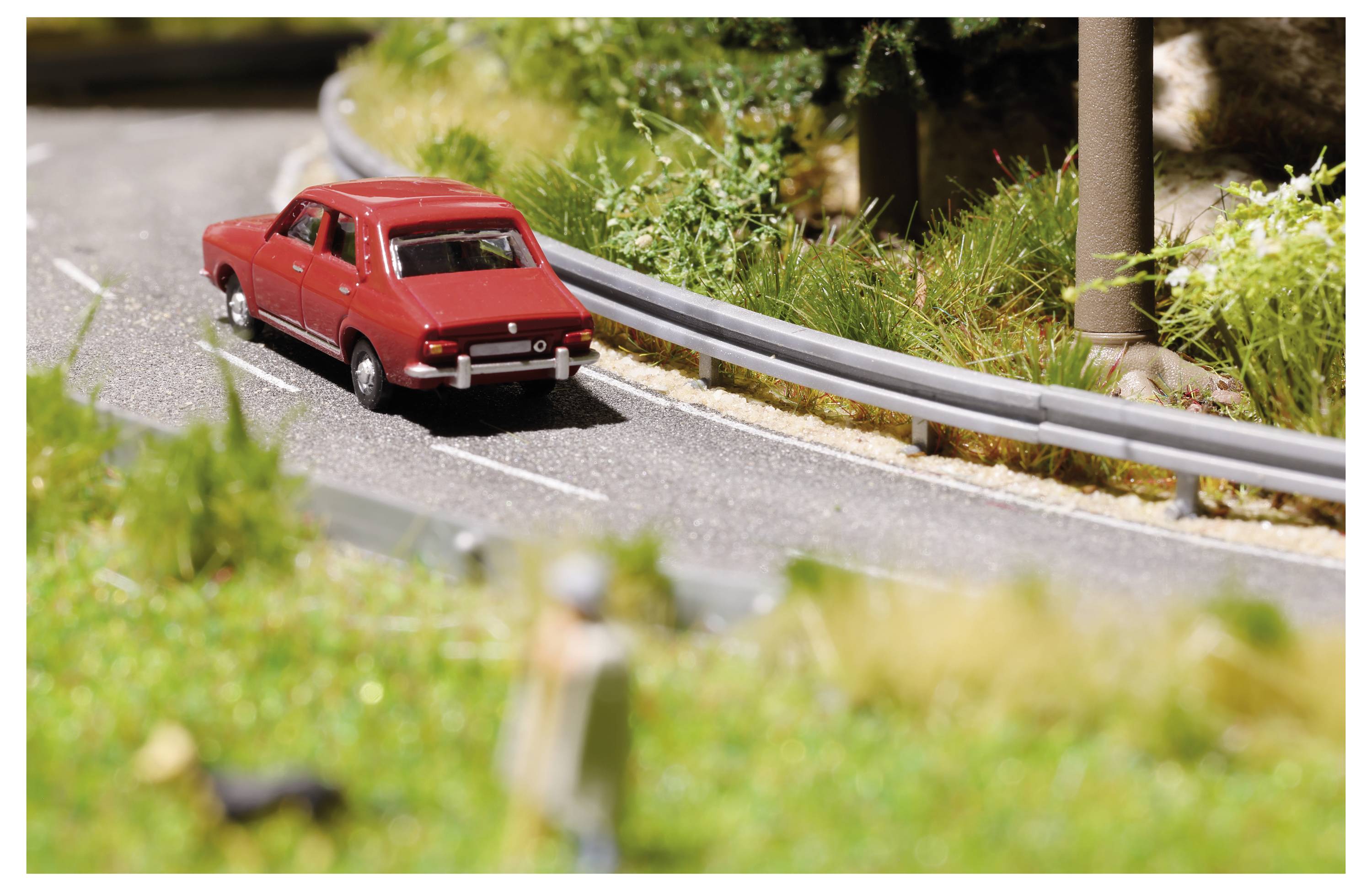 Rotes Spielzeugauto, das auf einer miniaturisierten, geschwungenen Straße neben einer Schutzplanke entlangfährt, mit verschwommenem Gras und Bäumen im Hintergrund.