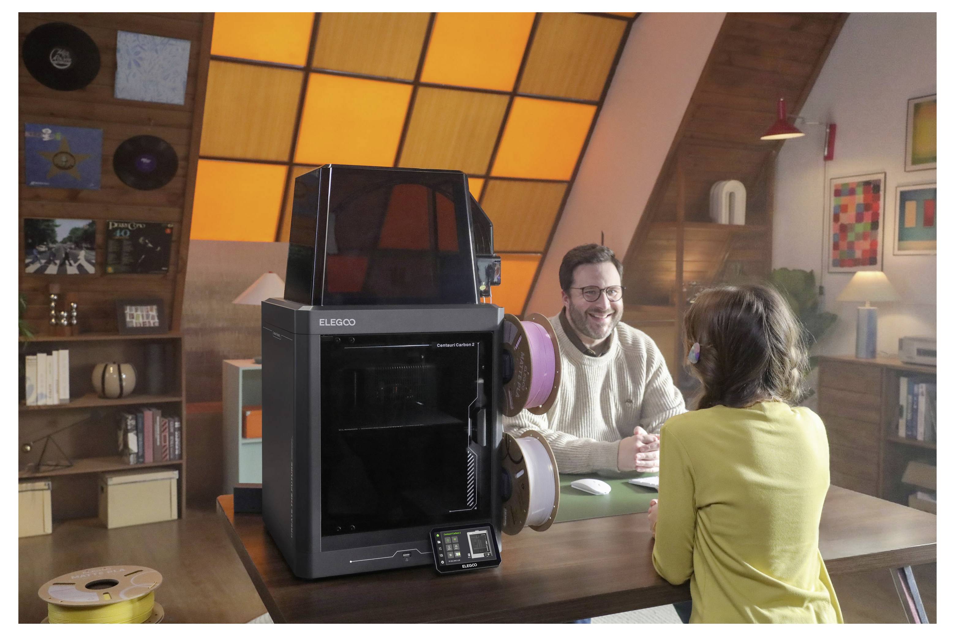 Zwei Personen unterhalten sich an einem Tisch in einem gemütlichen Studio mit einem großen 3D-Drucker im Vordergrund.