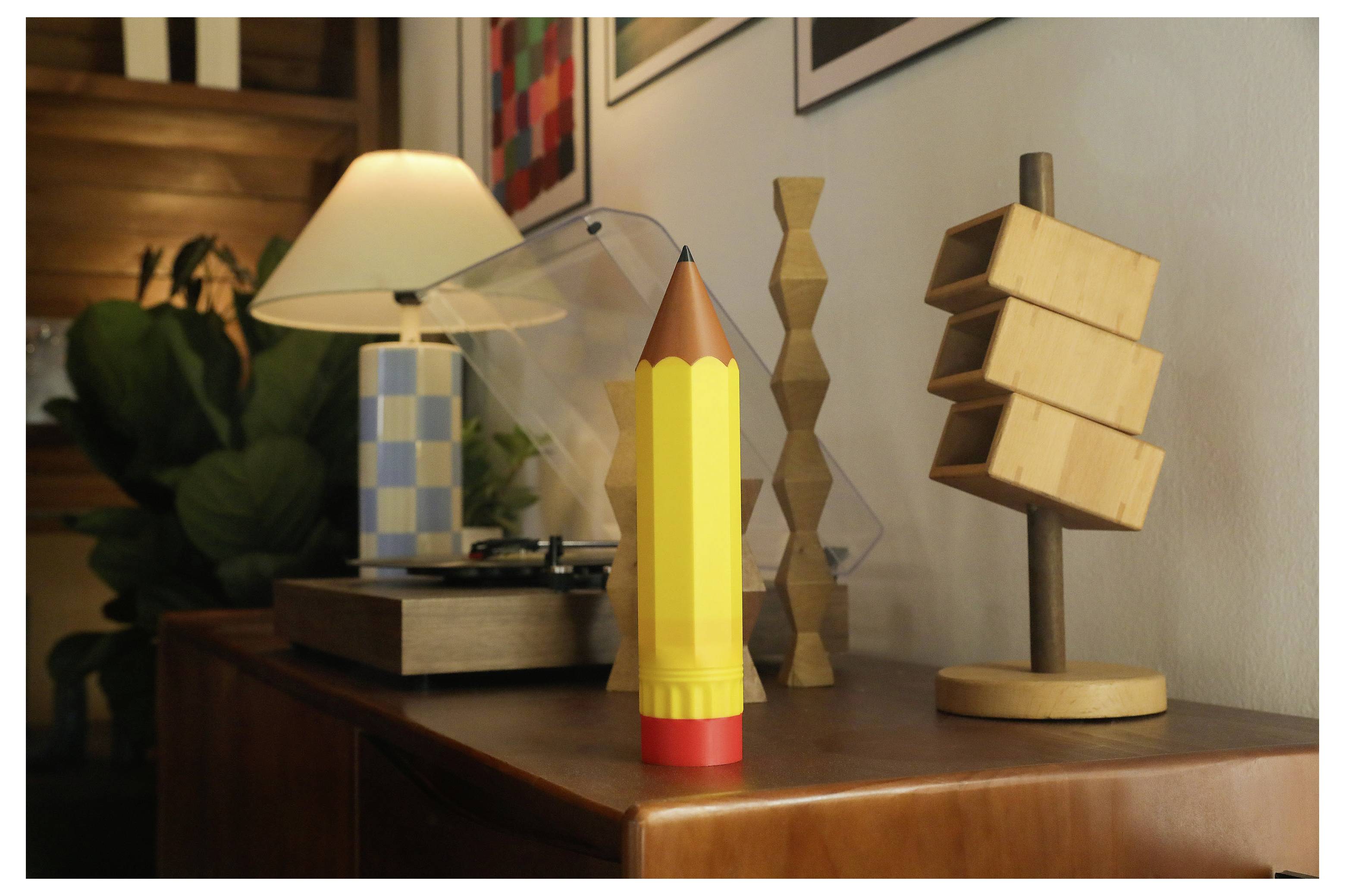 Ein großer gelber Bleistift steht auf einem Holztisch neben einer Lampe und kleinen Holzfiguren.