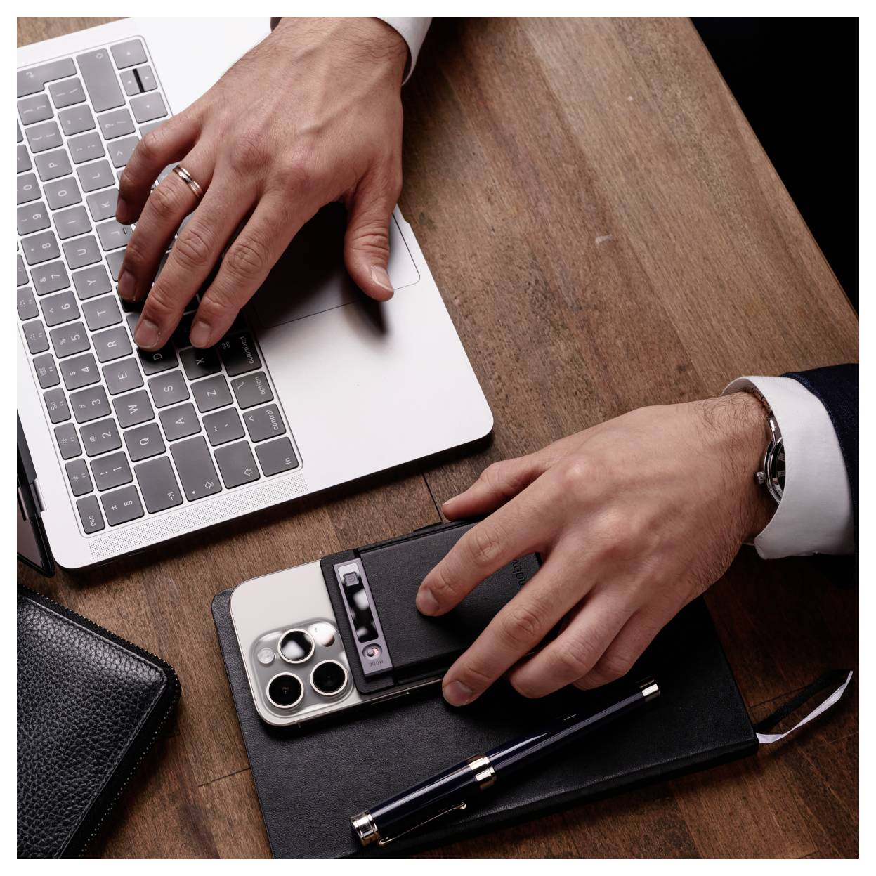 Hände, die einen Laptop und ein Smartphone auf einem Holzschreibtisch verwenden, mit einem Stift und Notizbuch in der Nähe, was auf Büroarbeit oder Multitasking hindeutet.