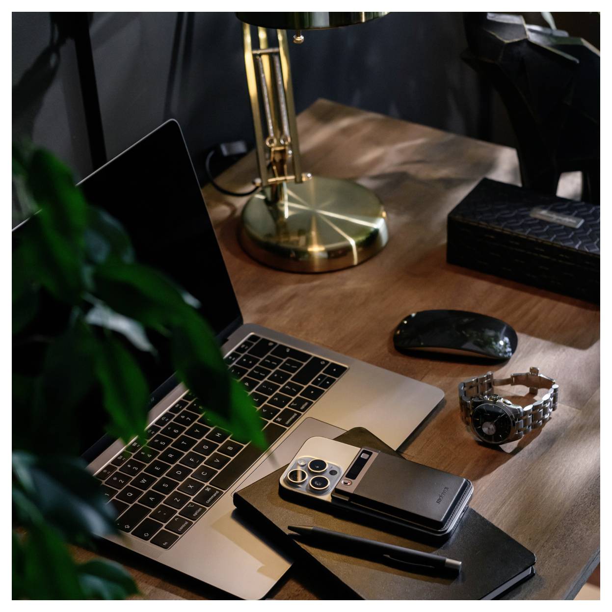 Ein Laptop auf einem Holzschreibtisch mit einem Smartphone, einer Smartwatch, einer Maus, einem Notizbuch und einem Stift neben einer Messingschreibtischlampe.