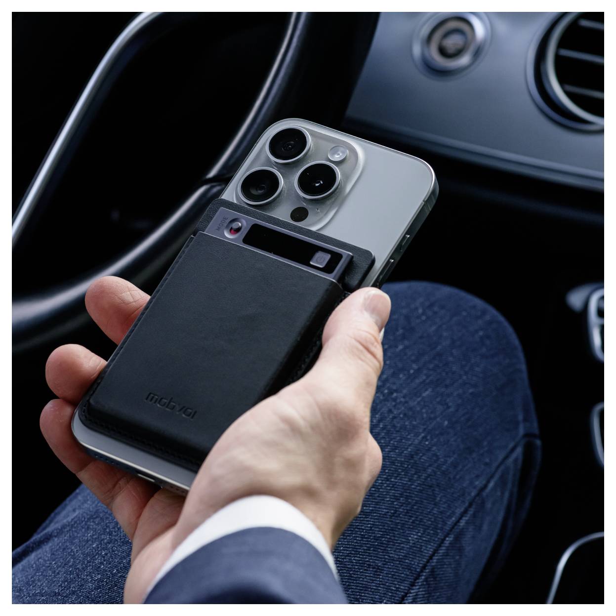 Hand hält ein Smartphone in einem Auto, mit einer schwarzen MagSafe-ähnlichen Powerbank auf der Rückseite befestigt.