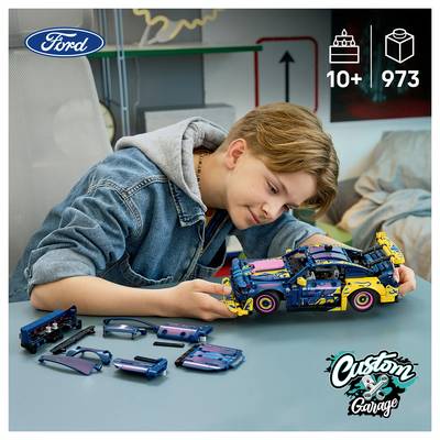 Junge, der ein LEGO-ähnliches Ford-Automodell an einem Tisch zusammenbaut, mit zerlegten Teilen neben ihm und „10+" und „973" in der Ecke angezeigt.
