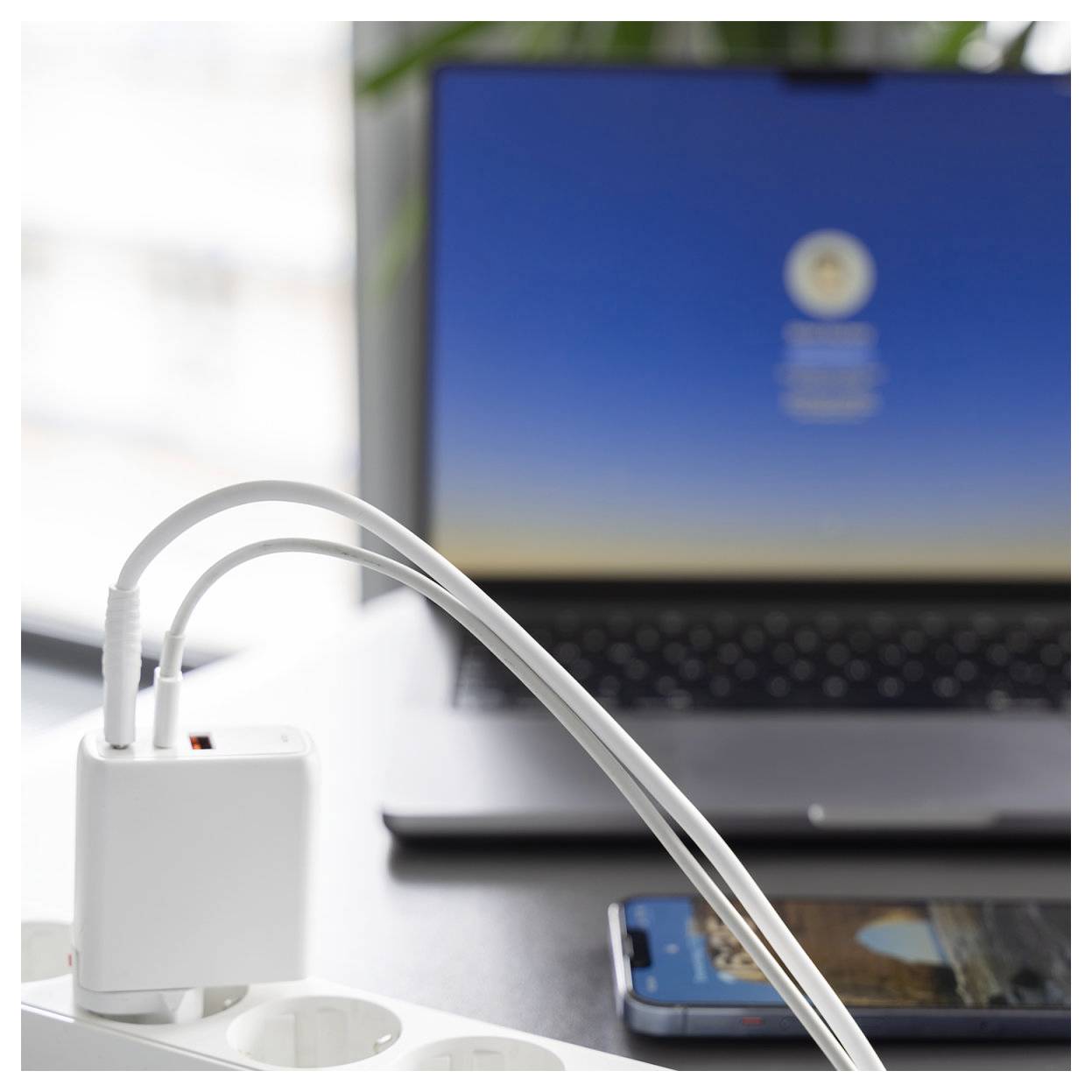 Laptop und Smartphone werden von einem weißen Netzteil auf einem Schreibtisch aufgeladen, wobei Kabel in eine Steckdosenleiste im Vordergrund eingesteckt sind.