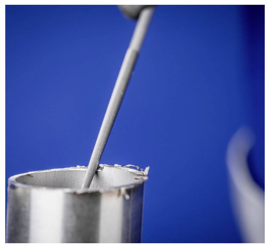 Nahaufnahme eines Metallstabs, der in einem kleinen silbernen Becher vor blauem Hintergrund umrührt.