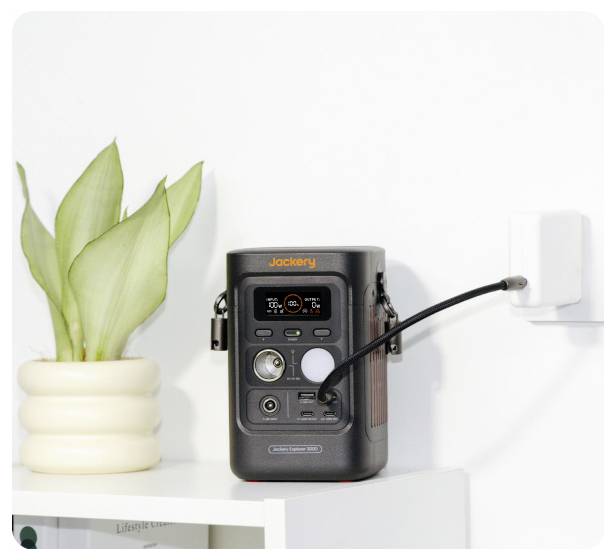 Jackery tragbare Stromstation, die in eine Wandsteckdose neben einer Topfpflanze auf einem weißen Regal eingesteckt ist.