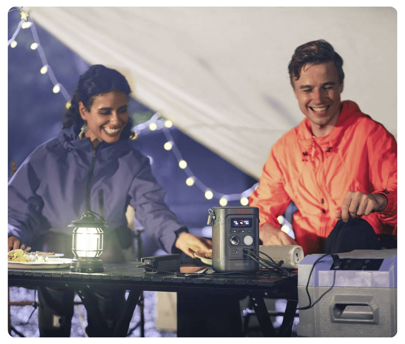 Zwei Personen in Regenjacken lächeln, während sie eine Campingmahlzeit an einem Tisch mit einer Laterne, einer tragbaren Stromstation und einer Kühlbox in einem Zelt zubereiten.