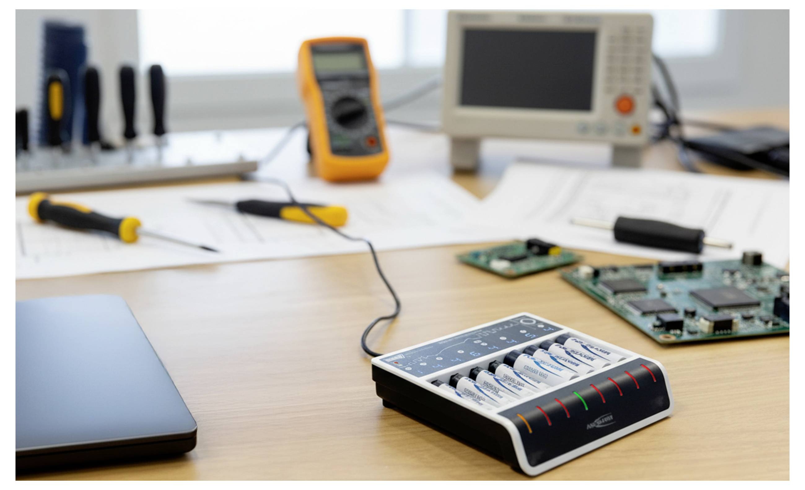 Elektronische Reparaturwerkbank mit einem Batterielader, Leiterplatten, Werkzeugen, einem Laptop und Prüfgeräten auf einem Schreibtisch.