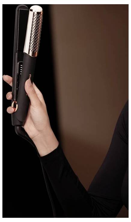 Hand hält ein schwarzes Haarglättungsgerät mit silbernen belüfteten Platten vor einem dunklen Hintergrund.