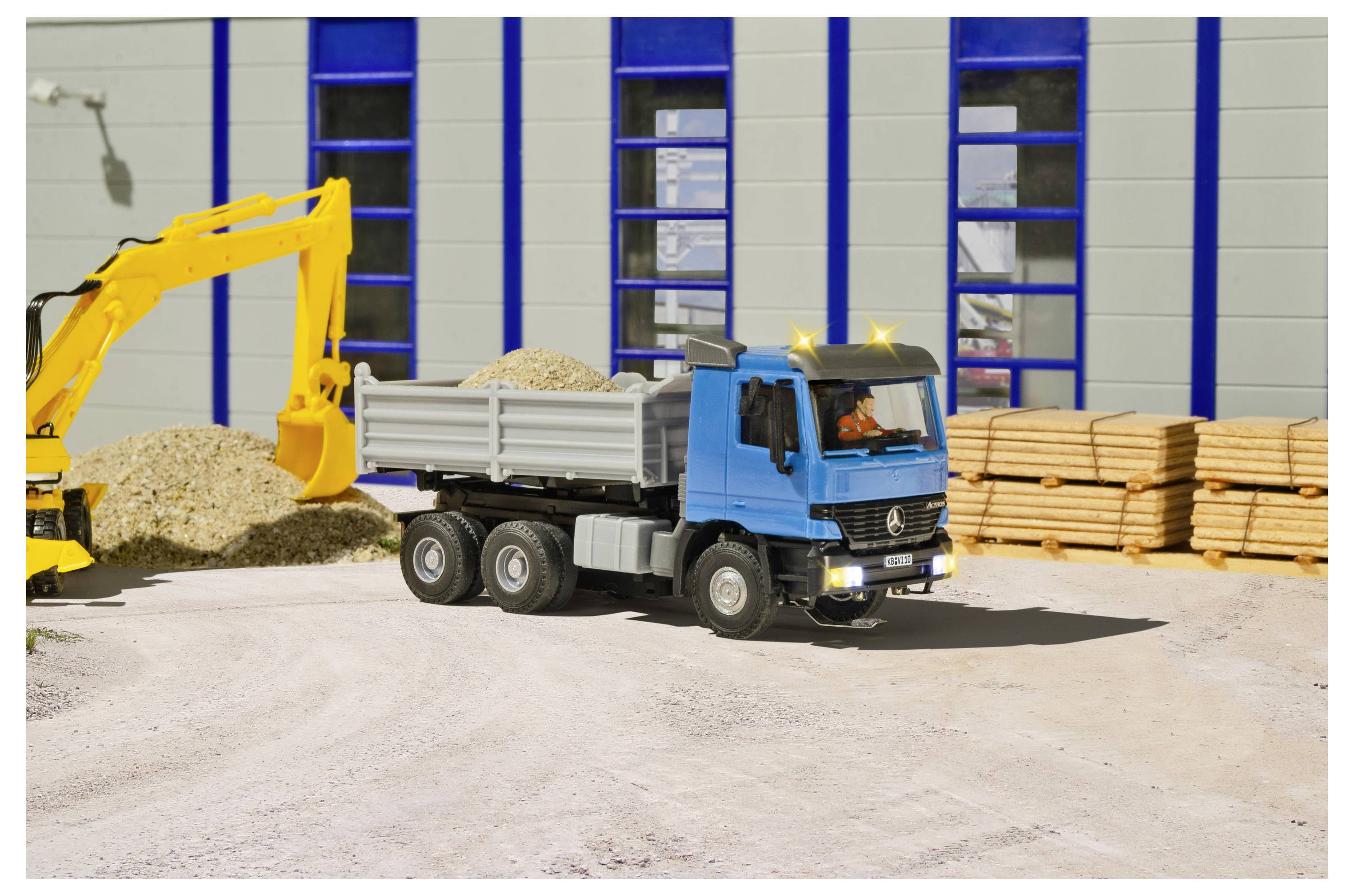 Blauer Kipplaster wird von einem gelben Bagger auf einer Baustelle beladen, im Hintergrund Holzstapel und ein Lagerhaus.