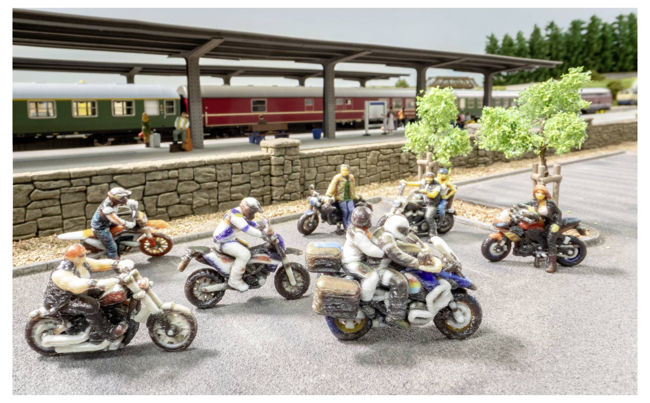 Miniaturszene mehrerer Motorradfahrer, die auf einer befestigten Straße neben einem Bahnsteig mit abgestellten Zügen und Bäumen fahren.