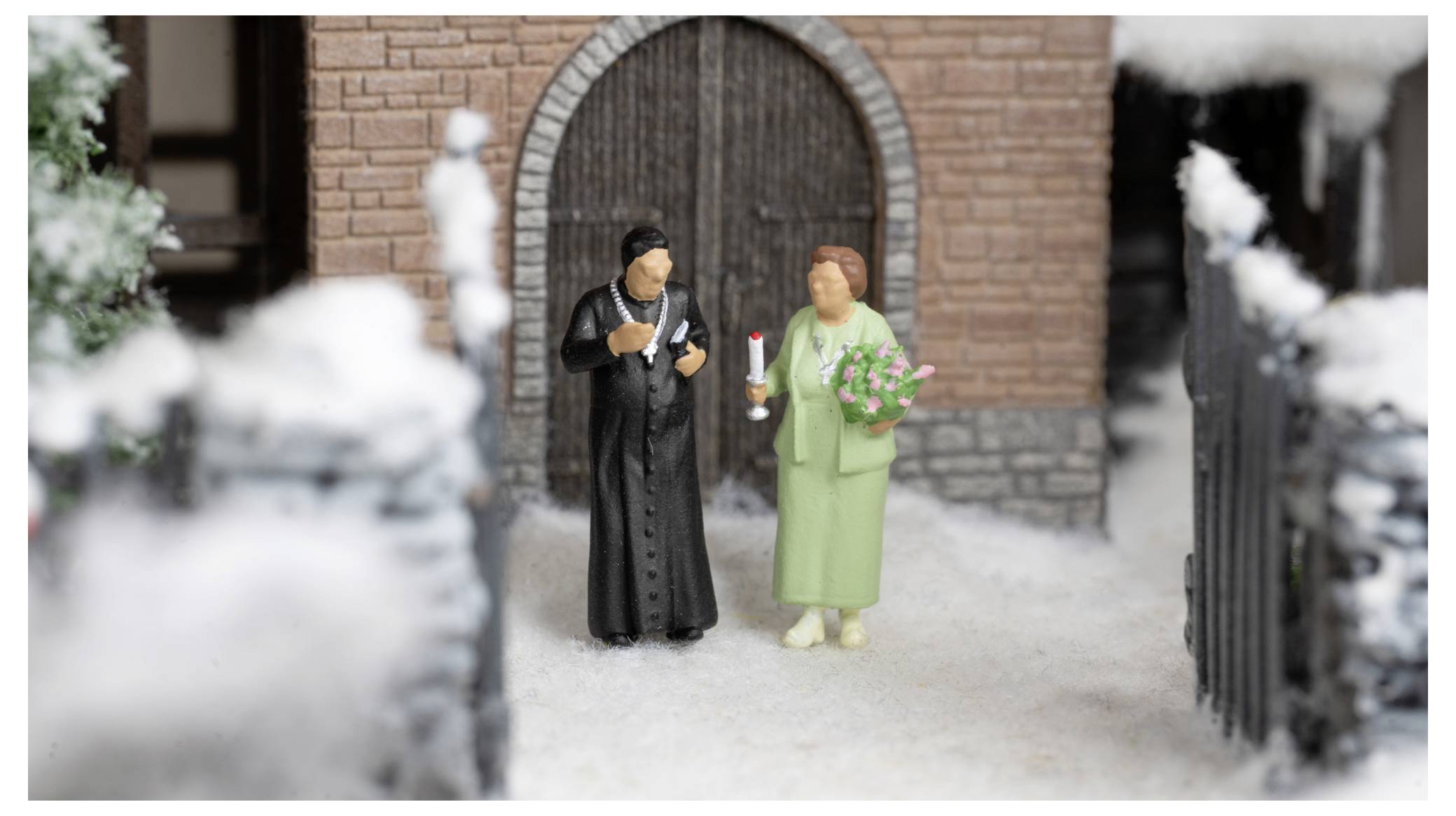 Miniatur-Braut- und Bräutigam-Figuren stehen vor einem Backsteingebäude, gekleidet in Schwarz und Grün, und halten Blumen und eine Kerze in den Händen.