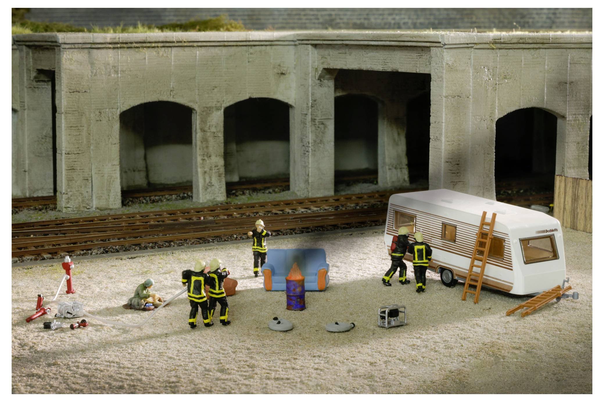 Modellzugszene mit Feuerwehrleuten, die auf einen kleinen Brand neben einem Wohnwagen in der Nähe von Eisenbahngleisen und einem Tunneleingang reagieren.
