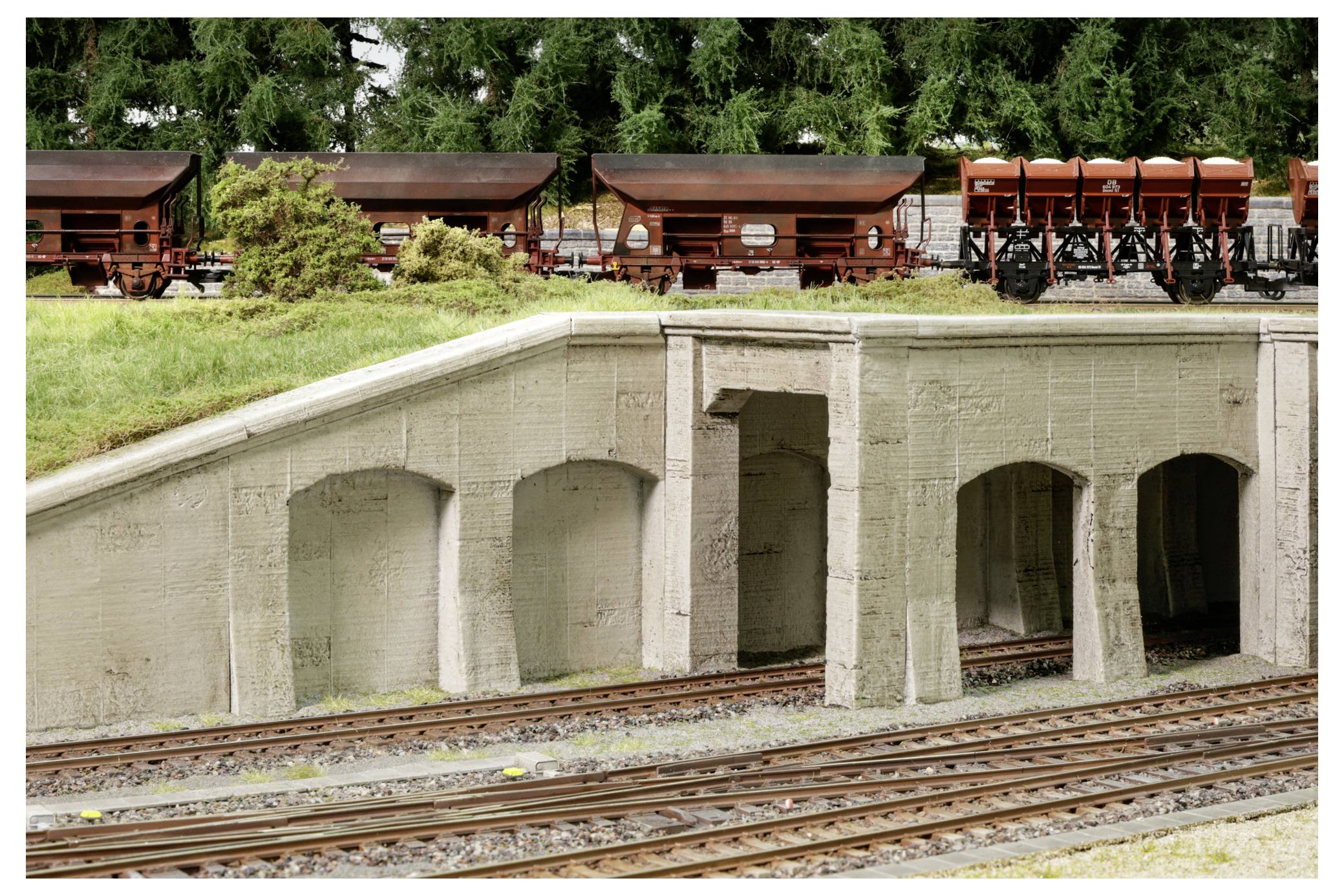 Modelleisenbahnszene mit einer Betontunnel-/Unterführungsstruktur neben mehreren Gleisen und abgestellten Güterwagen auf einem grasbewachsenen Damm.