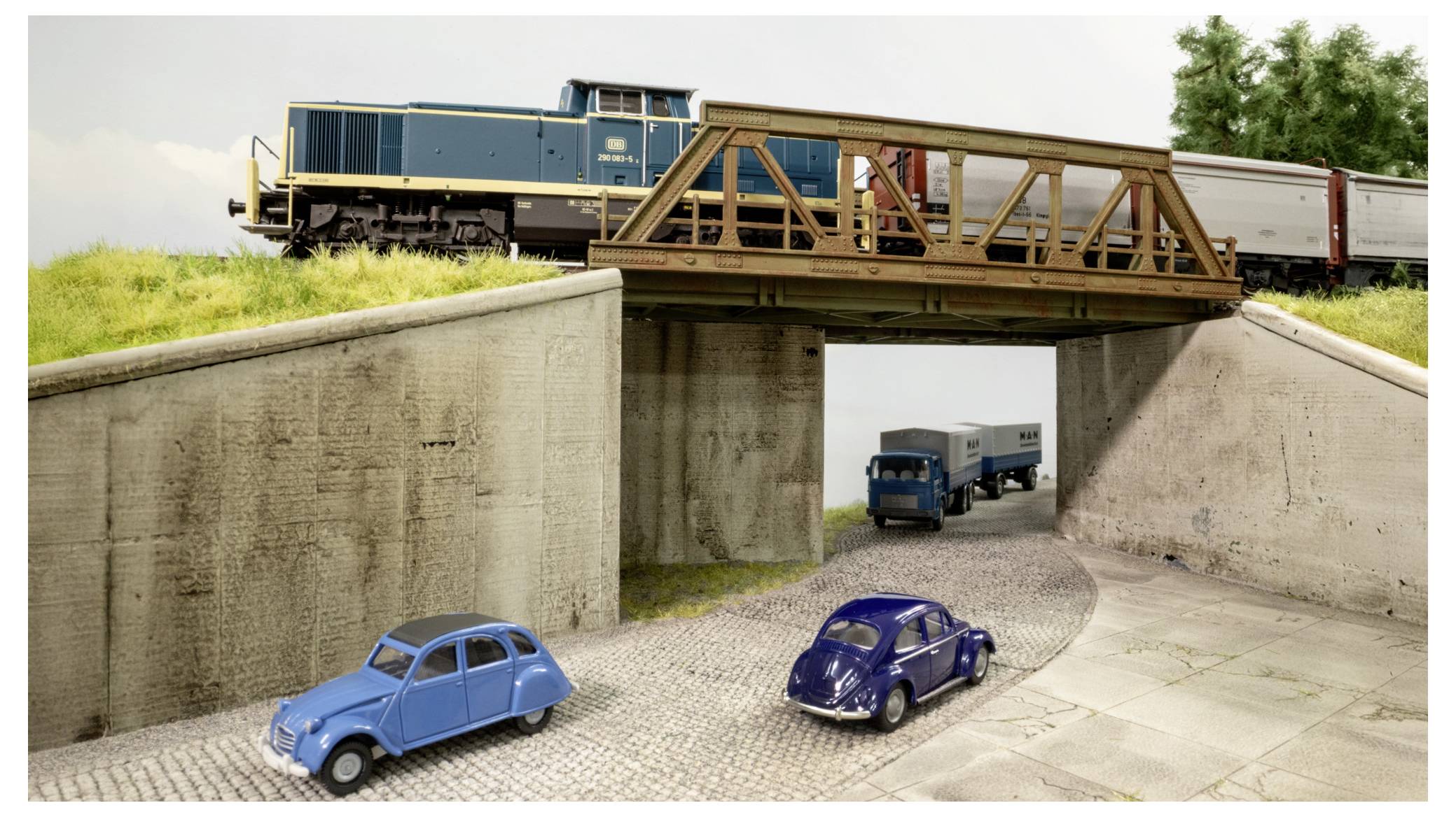 Ein Spielzeugeisenbahn überquert eine Brücke über eine Straßenunterführung, während zwei Spielzeugautos unten auf dem Gehweg stehen.