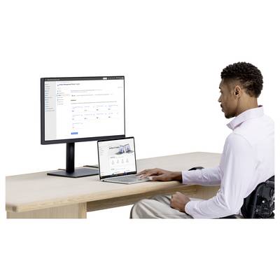 Ein Mann im Rollstuhl nutzt einen Laptop neben einem externen Monitor, der ein Website-Dashboard anzeigt, während er an einem Schreibtisch arbeitet.